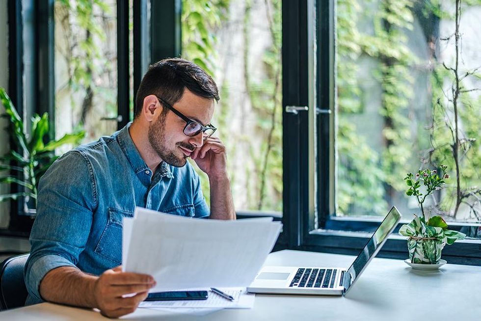 Un hombre con papeles está leyendo algo en su computadora portátil.