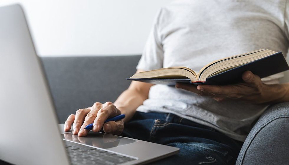 Man reads a book while working on his laptop