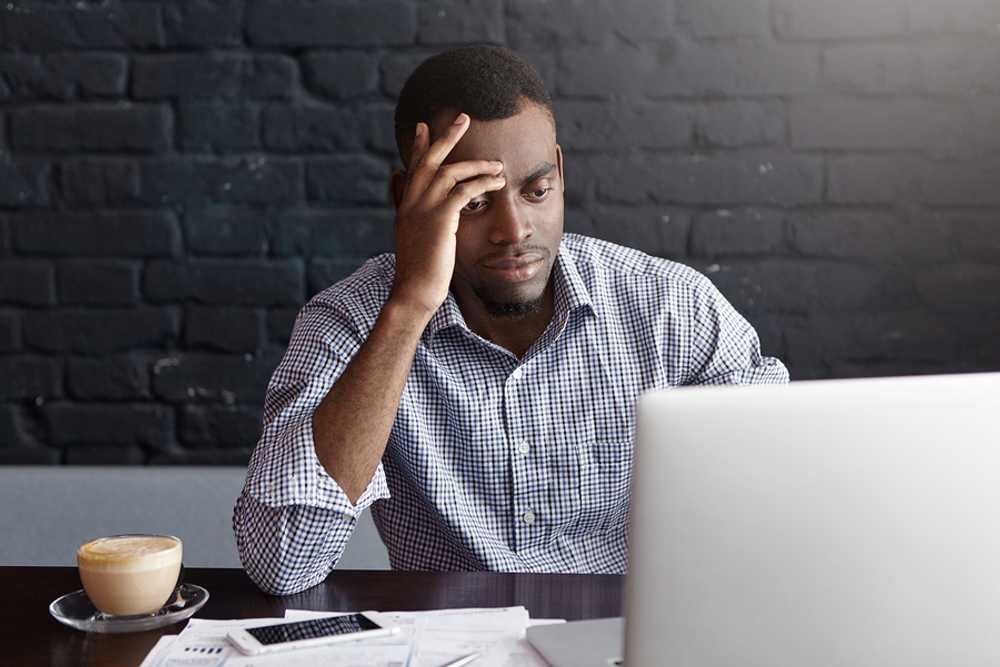 A stressed young professional sits at a laptop with his hand on his forehead, looking discouraged after receiving a job rejection, with papers and a coffee cup on the desk