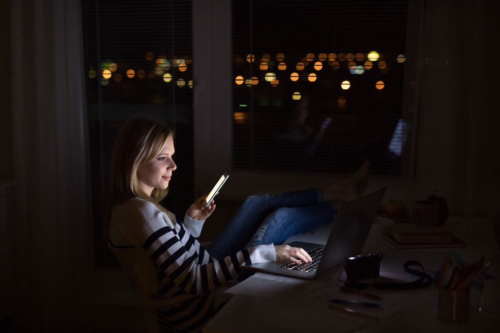 Woman at desk, holding smartphone, working on laptop at night.