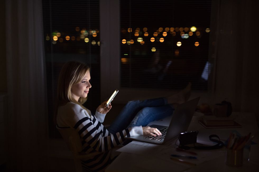 Woman at desk, holding smartphone, working on laptop at night