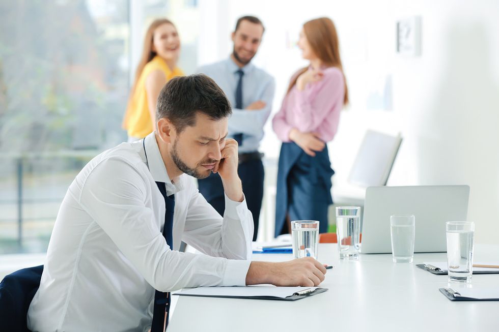 Young man looking sad at his desk as his coworkers laugh and talk behind him.