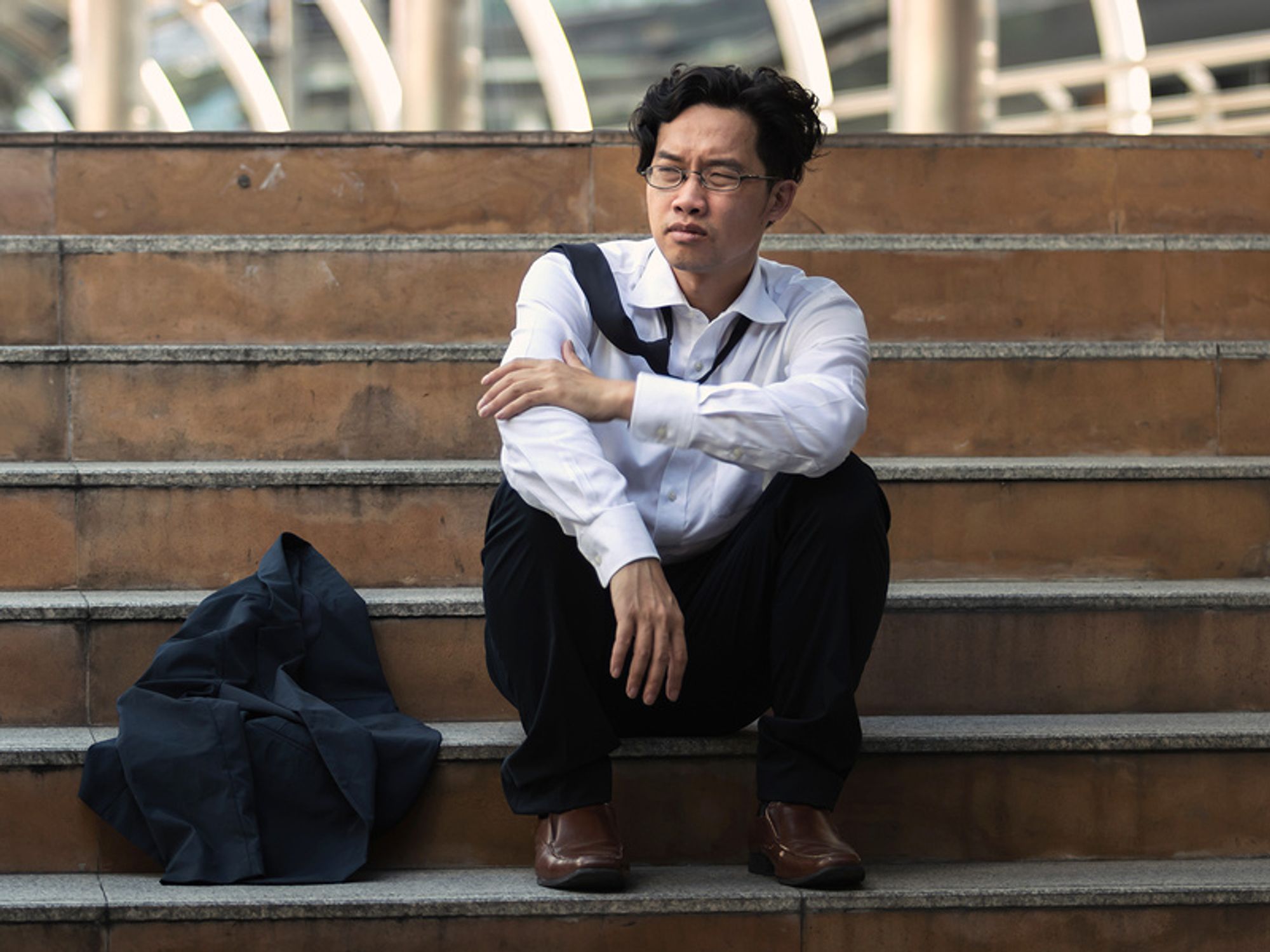 Young professional man sitting on steps, feeling upset that he's unemployed.