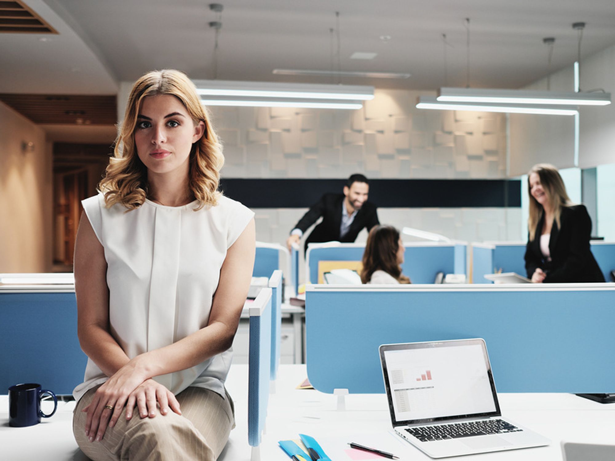 Young woman sitting alone at a cubical while her co-workers laugh and talk in the background.
